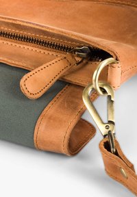 Braune Ledertasche mit einem grünen Canvas-Abschnitt, ausgestattet mit einem Reißverschluss, Messingbeschlägen und gesteppten Details an den Kanten.