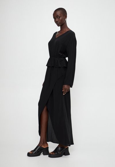 Black maxi dress with a V-neck, long sleeves, peplum waist, and side slit. Features a flowing fabric and stylish heeled sandals.