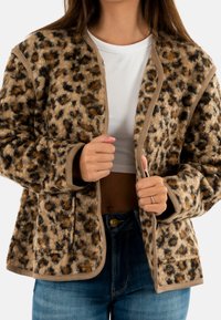 Donna che indossa una giacca leopardata pelosa sopra un top corto bianco e jeans blu, tenendo le estremità aperte della giacca.