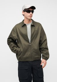 Νέος άνδρας με γκρι καπέλο, μαύρα γυαλιά ηλίου, λαδί ζακέτα με φερμουάρ, ανοιχτό γκρι μπλουζάκι και μαύρα cargo σορτς σε λευκό φόντο.