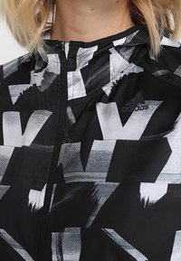 Gros plan sur une veste de sport Adidas noire et blanche avec un motif abstrait et une fermeture éclair à l'avant, portée par une personne aux cheveux blonds.
