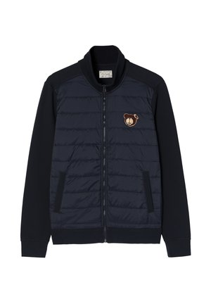 Tumši zilas krāsas jaka ar quiltētām priekšējām paneļiem, ribotām apkaklēm un adītām piedurknēm. Uz krūtīm ir lāča emblem, kā arī ir rāvējslēdzēja kabatas.