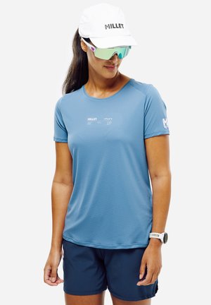 Frau trägt ein blaues Kurzarmshirt, marineblaue Shorts, weiße Sportkappe, reflektierende Sonnenbrille und weiße Armbanduhr, steht vor einem einfarbigen Hintergrund.