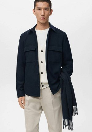 Camisa azul marino de botones con dos bolsillos en el pecho, combinada con un cárdigan crema y pantalones gris claro, sostenida con una bufanda oscura colgada del brazo.