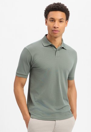 Man met krullend haar, gekleed in een donkergroen poloshirt met korte mouwen en beige broek, staand met handen in de zakken tegen een witte achtergrond.