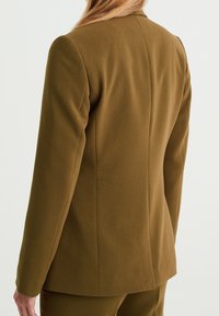 Hnědý saka s přizpůsobeným střihem, strukturovaným designem, hladkou texturou a dlouhými rukávy. Obsahuje jeden zadní šev a minimální detaily.
