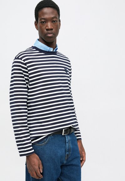 Navy and white striped long-sleeve shirt with a ribbed crew neckline and a small logo on the chest, paired with blue denim jeans and a black belt.