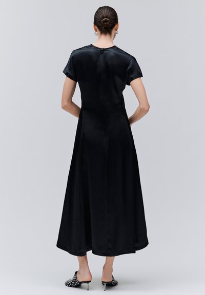 Femme aux cheveux attachés en chignon portant une robe midi noire à manches courtes et des mules à talons cloutées noires, debout face à un fond gris.