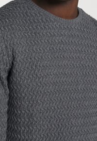 Gros plan sur une personne à la peau foncée portant un pull tricoté gris texturé avec un motif tissé et un col rond.