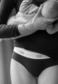 Schwarze Unterwäsche mit einem weißen elastischen Bund, der das "cache cœur"-Logo trägt. Ein weicher grauer Pullover ist um ein Baby gewickelt, das in einem Arm gehalten wird.