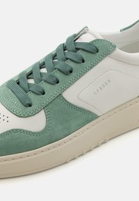 Grüne Wildleder- und weiße Leder-Sneaker mit strukturierten Akzenten, runder Zehenpartie, flachem beigen Sohlen und tonalen Schnürsenkeln. Minimale Markenpräsenz an der Seite.