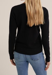 Maglione nero testurizzato con maniche lunghe e bordo a costine, indossato con jeans in denim blu. La vista dal retro rivela una vestibilità rilassata e cuciture sottili.