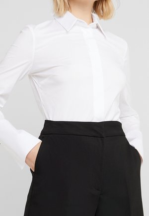 Persoon die een wit shirt met lange mouwen draagt, ingestopt in een zwarte hoge taille broek, met handen in de zakken.