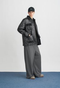 Młody mężczyzna ubrany w czarną pikowaną kurtkę, szare szerokie spodnie, beżowe buty i czapkę w kolorze czarnym, stojący przy białej ścianie na niebieskiej podłodze.