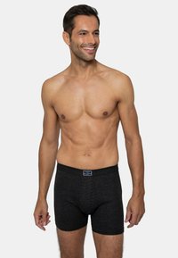 Schwarze Boxer-Shorts aus strukturiertem Stoff mit elastischem Bund, getragen von einem Mann, der vor einem schlichten Hintergrund steht.