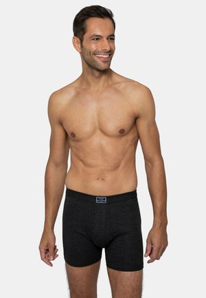 Schwarze Boxer-Shorts aus strukturiertem Stoff mit elastischem Bund, getragen von einem Mann, der vor einem schlichten Hintergrund steht.