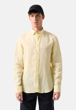 Young man wearing a pale yellow button-up shirt and a black baseball cap, standing against a plain light background.