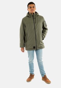 Giacca impermeabile verde oliva con cappuccio, chiusura lampo frontale, due tasche laterali e una tasca sulla manica. Indossata con jeans azzurri e scarpe marroni.