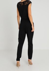 Black jumpsuit featuring a lace-accented neckline, fitted waist, and tapered legs, paired with beige high heels.