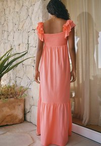 Robe maxi en corail avec des accents de volants sur les épaules, un bodice smocké et une jupe à volants fluide, devant un mur en pierre et un jardin.