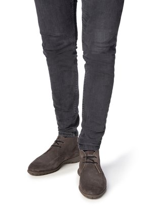 Jambes portant un jean gris foncé ajusté et des bottines en daim brun foncé avec des lacets, posées sur un fond blanc.