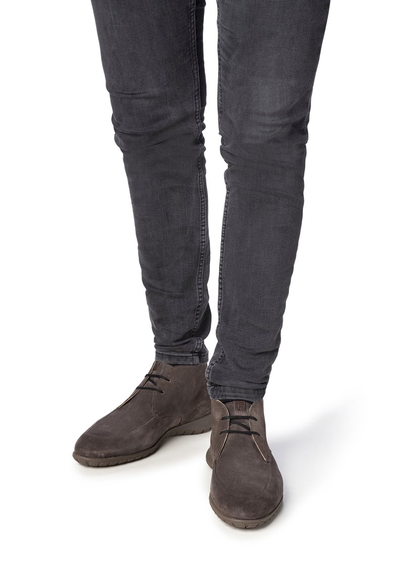 Jambes portant un jean gris foncé ajusté et des bottines en daim brun foncé avec des lacets, posées sur un fond blanc.
