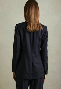 Person med glat, skulderlangt brunt hår iført en mørk tekstureret blazer og matchende bukser, set bagfra mod en ensfarvet lys baggrund.