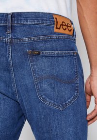 Jeans in denim blu con etichetta in pelle che mostra "Lee." Presentano una tasca posteriore cucita e cuciture a contrasto. Dettaglio ravvicinato dell'area della cintura.