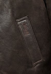 Barna bőrdzseki részlet gazdag textúrával, rajta varrott bőrcímke, amelyen sötétbarna betűkkel szerepel a „Schott NYC” felirat.