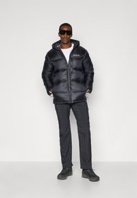 Schwarze wattierte Jacke mit Kapuze, ausgestattet mit einem Reißverschluss und einem Columbia-Logo. Getragen mit grauen Cargo-Hosen und schwarzen Wanderschuhen.
