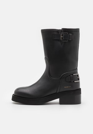 Bottillon en cuir noir avec un bout arrondi, un talon bloc, un détail de boucle sur le côté et une finition texturée. Présente un branding subtil en doré.