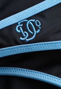 Gros plan sur un tissu noir avec des coutures bleu clair et des lettres entrelacées "ID" brodées en fil bleu clair.