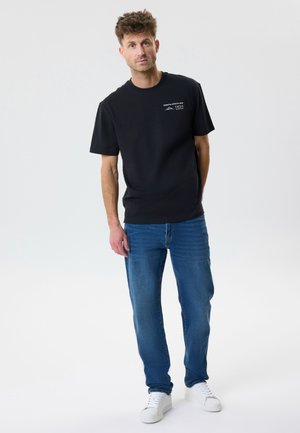 Man standing in black short-sleeve shirt with small white logo, blue jeans, and white sneakers against a plain light background.