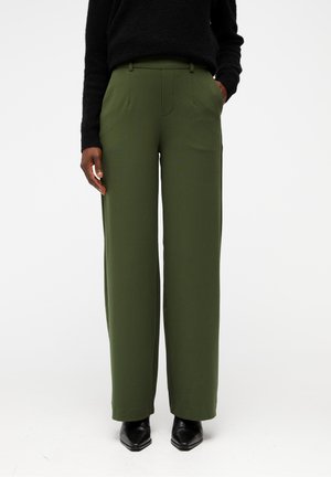 Personne portant un pantalon large kaki, un pull noir et des chaussures pointues noires, debout devant un fond blanc uni.