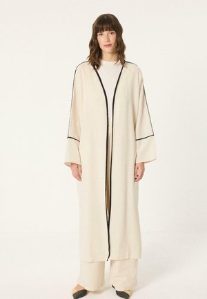 Kobieta ubrana w długi kremowy szlafrok z czarnym obszyciem na białą koszulę i spodnie, stojąca na jasnym, jednolitym tle.
