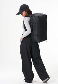 Schwarzer Rucksack aus strapazierfähigem Material, rechteckige Form mit Logo, mehrere Riemen und ein sicherer Verschluss. Wird von einer Person in sportlicher Kleidung getragen.