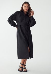Schwarzes langärmliges Hemdkleid mit Kragen, vorderen Knöpfen und tailliertem Gürtel. Mit einem seitlichen Schlitz und glatter Stofftextur, kombiniert mit schwarzen Sandalen.