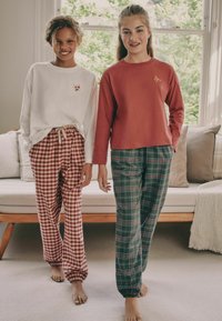 Zwei Modelle tragen Freizeitkleidung: ein weißes Langarmshirt mit floraler Stickerei, kombiniert mit rot karierten Hosen, und ein rostfarbenes Langarmtop mit grünem Karomuster.