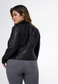 Veste en cuir noir avec un dos matelassé, dotée d'une finition lisse, de épaules structurées et d'un col montant, associée à un jean gris.