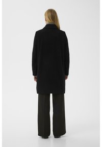 Schwarzer Mantel mit einem langen, geraden Design und großem Kragen. Getragen über grauen Streifen, kombiniert mit lockeren, dunklen Hosen. Minimalistischer Stil.