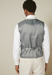 Gilet en satin gris avec une texture lisse, doté d'une boucle et d'un réglage par sangle à l'arrière et portant une chemise blanche à col en dessous.