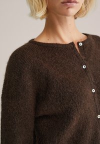 Cardigan tricotat maro featuring un material moale, texturat, guler rotund și cinci nasturi albi pe partea din față.