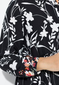 Abito nero con motivi floreali bianchi e foglie verdi, vita elastica e accenti floreali colorati sui polsini delle maniche.