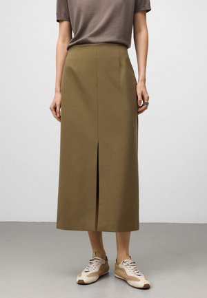 Donna con una gonna midi marrone con uno spacco frontale, sneakers beige e bianche, e un top marrone a maniche corte che si trova su un pavimento grigio.