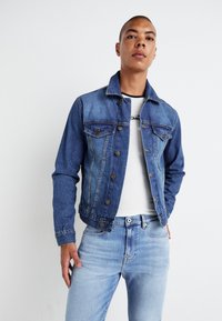 Mørkeblå denimjakke med knappelukking, to brystlommer og klassisk krage. Båret over en hvit T-skjorte.