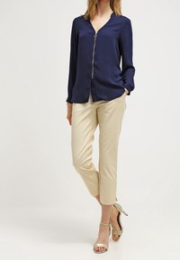 Námořnicky modrá blůza se zipem vpředu, dlouhými rukávy a hladkým povrchem, spárovaná s béžovými zkrácenými kalhotami a světlými zelenými sandály na podpatku.