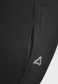 Schwarze Sportleggings mit einem Reißverschlusstasche und einem reflektierenden Logodetail an der unteren Seite. Weiche, glatte Textur.