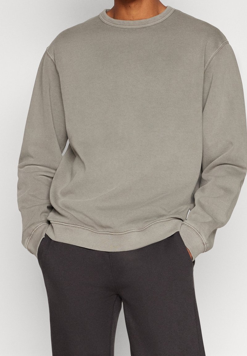 Homme portant un sweat-shirt ample gris clair à col rond et un pantalon gris foncé, les mains dans les poches, sur un fond uni.