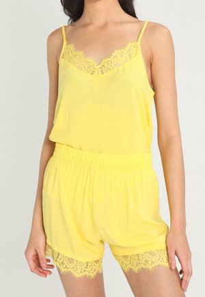 Vrouw draagt gele mouwloze cami met kanten rand en bijpassende shorts met elastische taille op een effen achtergrond.