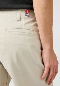 Beige bomuldsbukser med en baglomme, der har en trekantet stikning. Wrangler-label på linningen, subtil tekstur og klassisk pasform.
